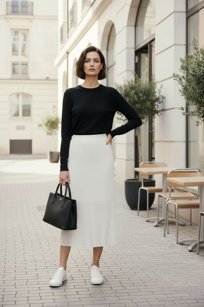 Woman in black top and white skirt standing on a city street with tables and chairs in the background.