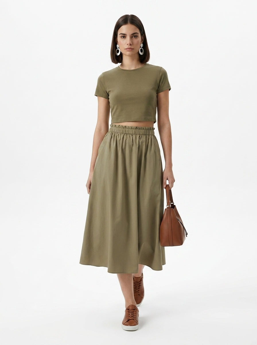 Woman wearing an olive green outfit with a skirt and top, holding a brown bag on a white background.