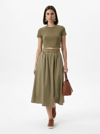 Woman wearing an olive green outfit with a skirt and top, holding a brown bag on a white background.