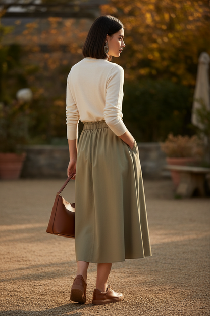 Woman in a cream sweater and green skirt holding a brown bag outdoors.
